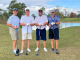 Four older men holding golf clubs on the course in front of a lake