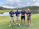 Four golfers wearing dark colored shirts holding their golf clubs in front of a lake
